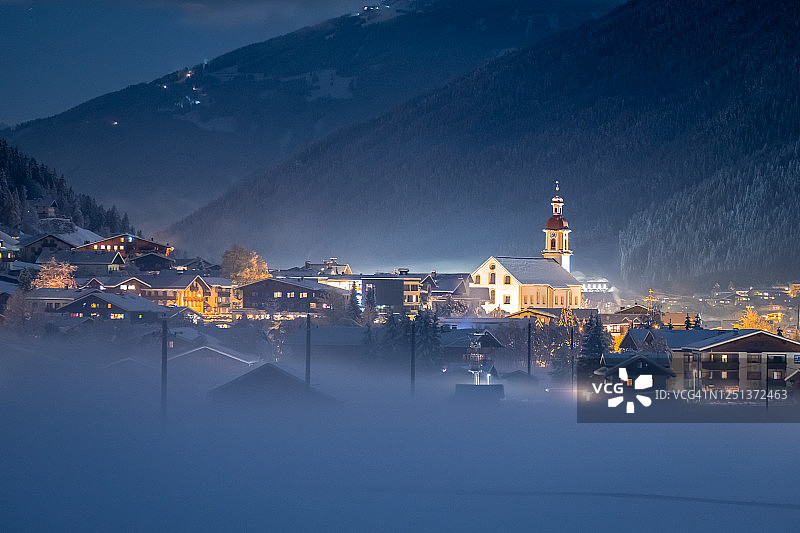 冬季夜景：俯瞰奥地利蒂罗尔州新施蒂夫特市和教堂，背景是山脉和云彩。山谷里有雾的寒冷夜晚图片素材