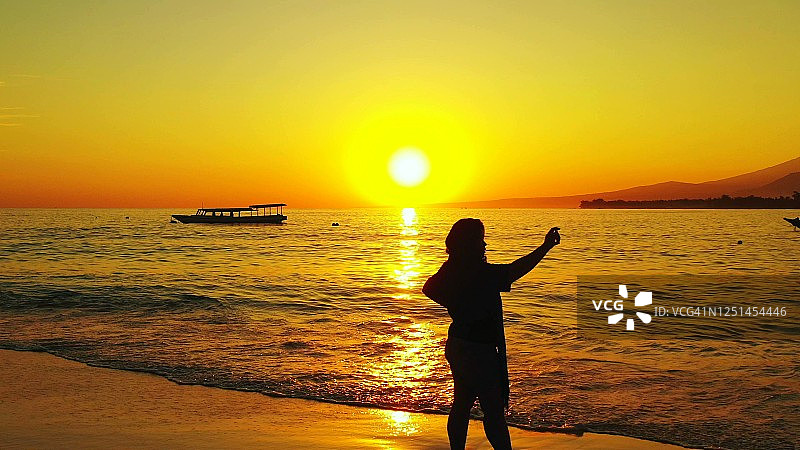 女游客带着旅行包在热带岛屿的岸边拍照，日落时分，黄色的太阳和天空倒映在停泊着船只的海面上图片素材