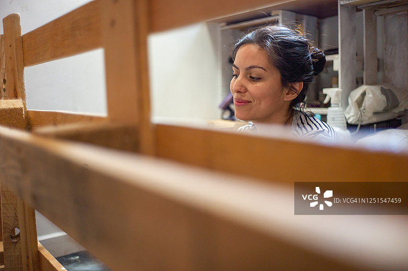 穿着黑白条纹长袖衬衫的年轻有魅力的西班牙裔女子在家笑着粉刷白色木托盘图片素材