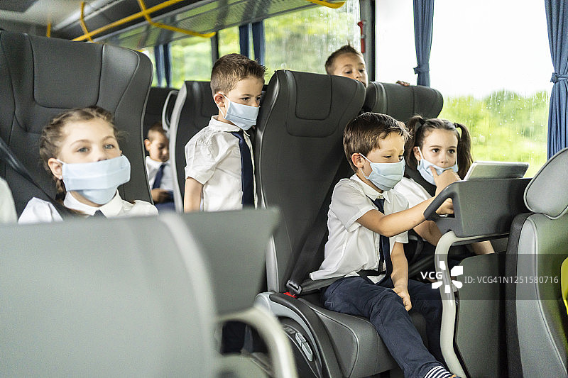 小学生坐在校车上，看着平板电脑并戴着口罩图片素材