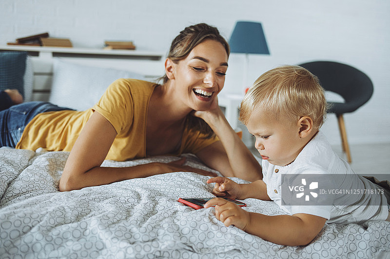 女人看着在床上玩智能手机的儿子图片素材