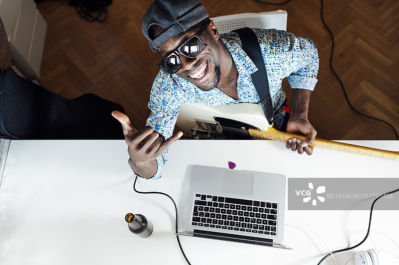 微笑的年轻男人坐在桌旁，拿着吉他和笔记本电脑，展示shaka手势图片素材