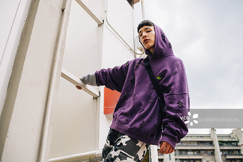 身穿连帽衫的酷炫青年站在金属梯子上，以天空为背景图片素材