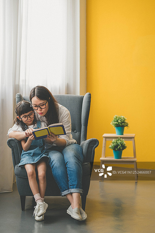快乐的年轻亚洲母亲和女儿在客厅沙发上阅读故事书图片素材