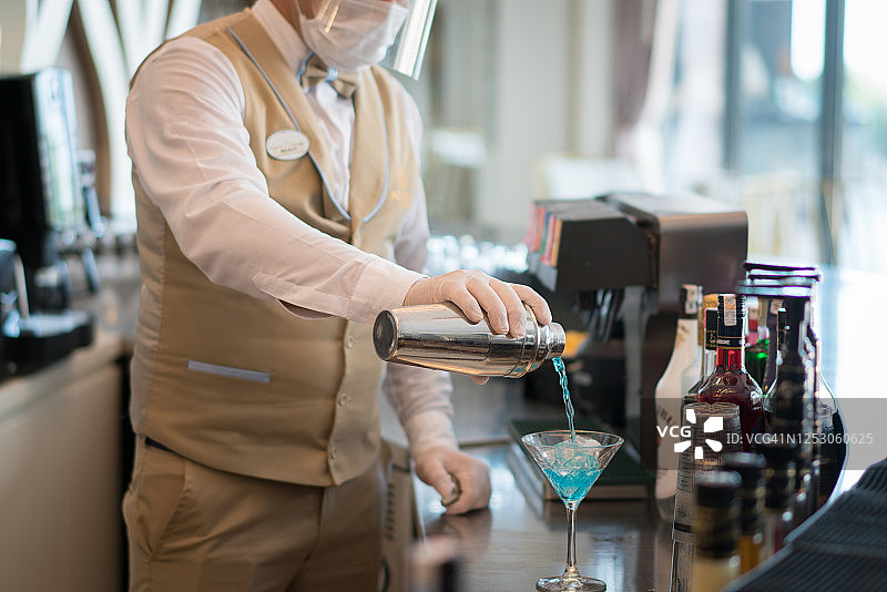 戴口罩的调酒师将蓝色液体从烧杯倒入装有冰块的玻璃杯中图片素材
