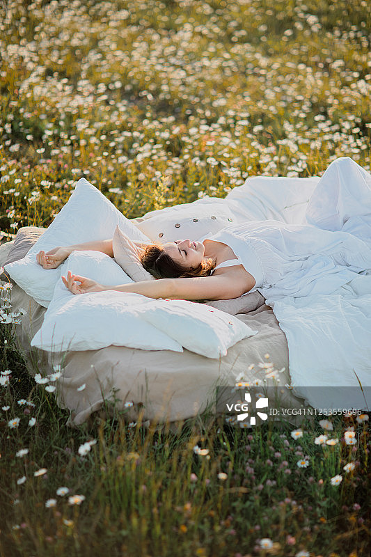 可爱的年轻卷发女人穿着白色连衣裙，躺在夏季洋甘菊田里柔软的亚麻床上。夏季假期灵感，良好睡眠和放松的概念图片素材
