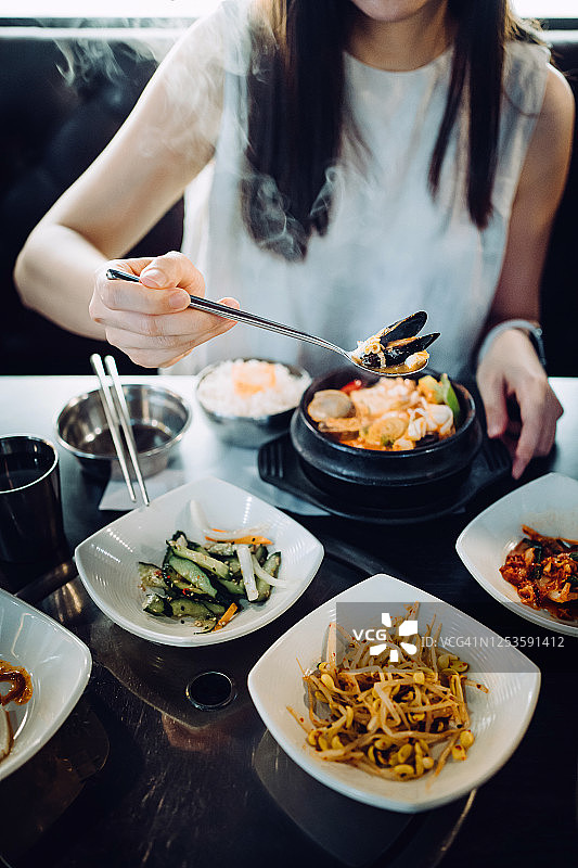 年轻亚裔女性在韩国餐厅享用韩式海鲜豆腐汤和各种小菜图片素材