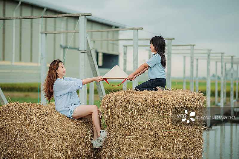 中国母亲坐在干草堆收割场上，将传统藤帽递给女儿图片素材