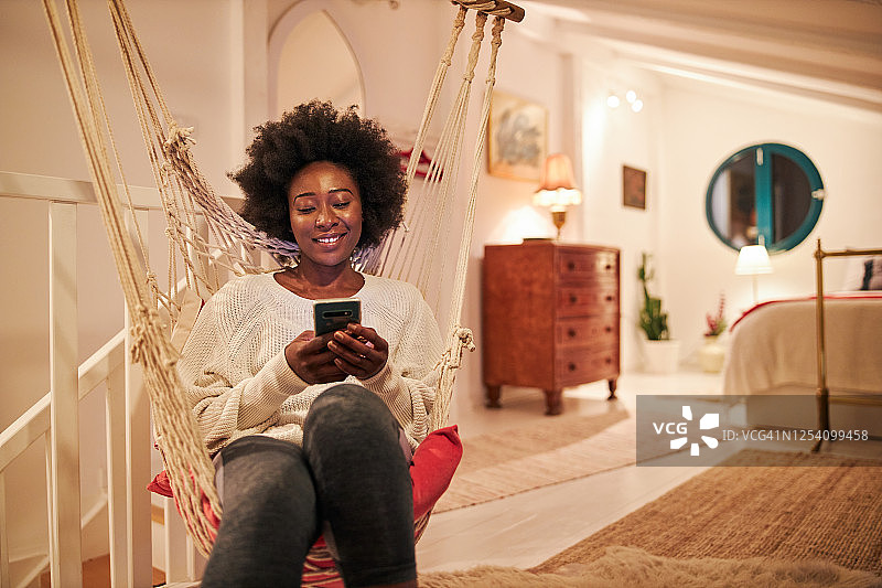 快乐的年轻女人在家中坐在秋千上使用手机图片素材