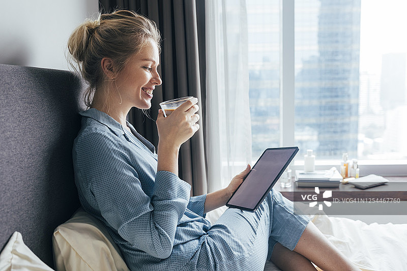 快乐的金发女子在早晨穿着蓝色睡衣在卧室里喝咖啡图片素材