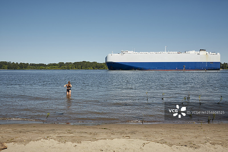 从索维岛看到的哥伦比亚河上行驶的大型运输车辆，一人站在水中显示比例图片素材