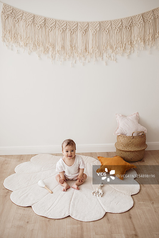 迷人的微笑幼儿坐在贝壳地毯上。可爱的宝宝与生态玩具，星枕头和棉花花边坐在美丽的温柔游戏室的地板上图片素材