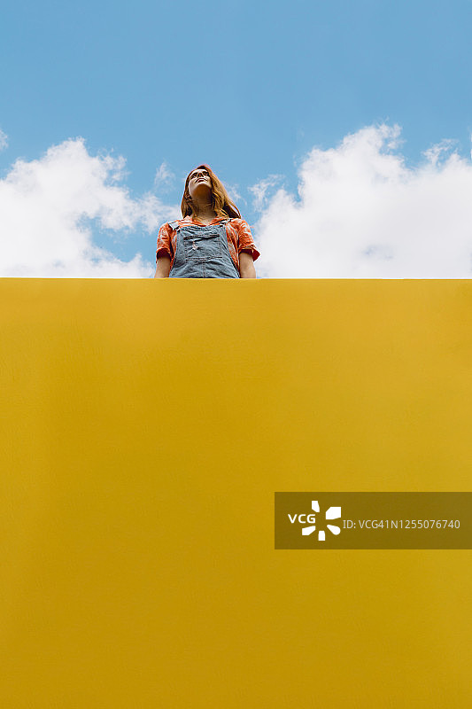 年轻女子站在多云天空下的黄色墙壁上方图片素材