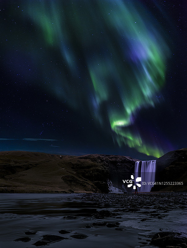 在冰岛中南部斯科加瀑布上方的北极光下伸开双臂的男人图片素材