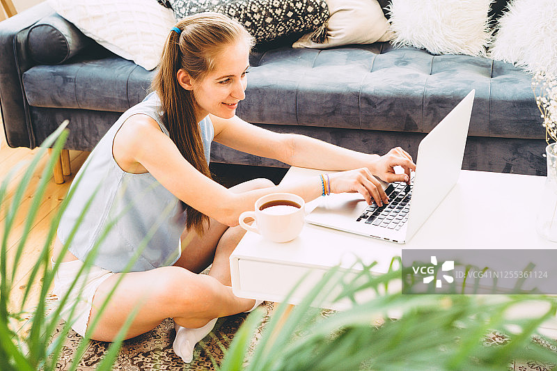年轻女子坐在笔记本电脑旁，穿着家居服的年轻女子在地板上用粉色马克杯喝茶图片素材