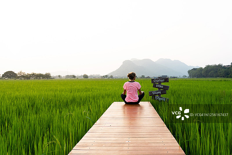 金发女子坐在泰国北碧府稻田中央的木制路尽头，旁边有路标图片素材