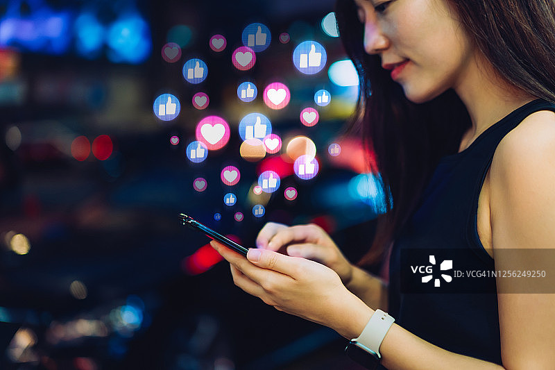 年轻漂亮的亚洲女性在社交媒体上使用智能手机，在城市夜晚浏览或点赞、喜欢、评论朋友和页面。社交媒体沉迷概念图片素材