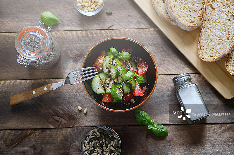 素食、纯素食和生食：西红柿、黄瓜、罗勒和绿色沙拉蔬菜沙拉，木桌上的坚果和种子，旁边是切片法棍图片素材