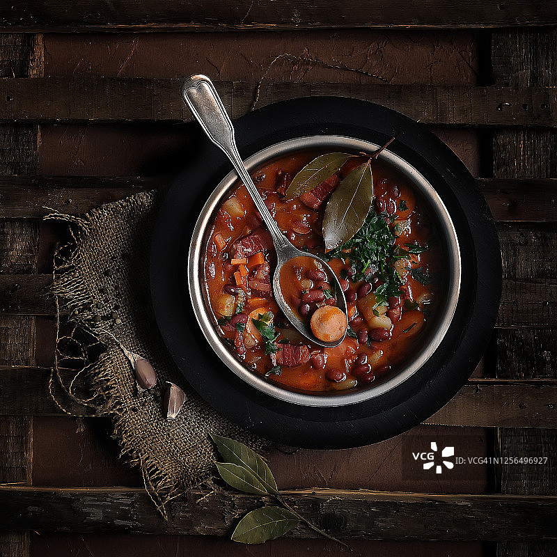 美味的红色传统德国番茄汤与肉制品、豆类、香草的食品摄影，顶部有织物和香料的棕色木质纹理背景上的老式勺子图片素材