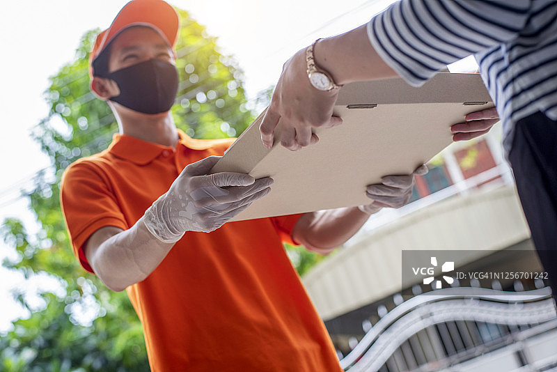 身穿绿色制服的快递员将纸板箱包裹交给顾客，双方均佩戴口罩以保护免受新冠病毒感染。新常态生活图片素材