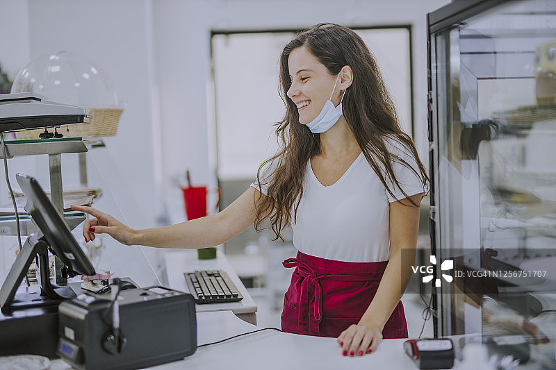 疫情期间，友好的年轻女收银员在咖啡店收银台后微笑，戴着防护口罩图片素材