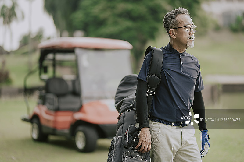 一位亚洲中年男子从高尔夫球车上拿起高尔夫球包，走向发球区准备开始比赛图片素材