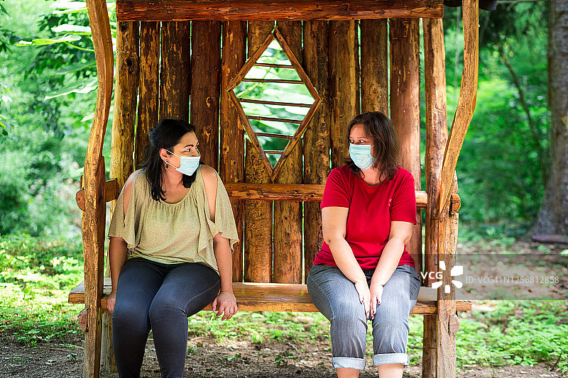 朋友们戴着口罩坐在公园长椅上，保持社交距离图片素材