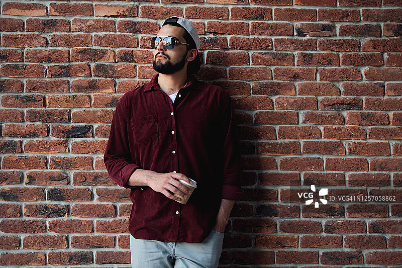 戴眼镜的严肃青年手持咖啡杯饮用，深色皮肤印度男子靠在砖墙上图片素材
