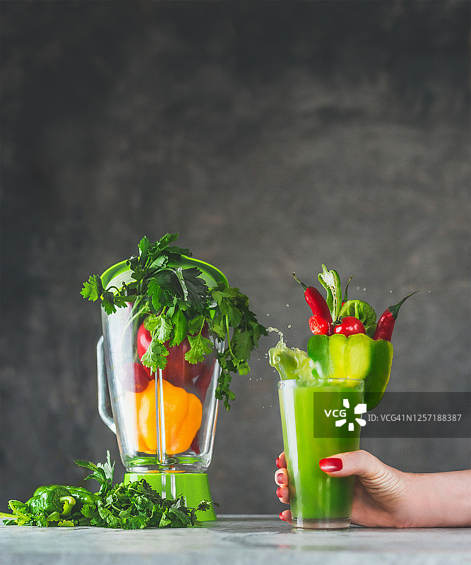 妇女手握盛有绿色蔬菜冰沙或果汁的玻璃杯，灰色桌子上放着搅拌机和蔬菜原料图片素材