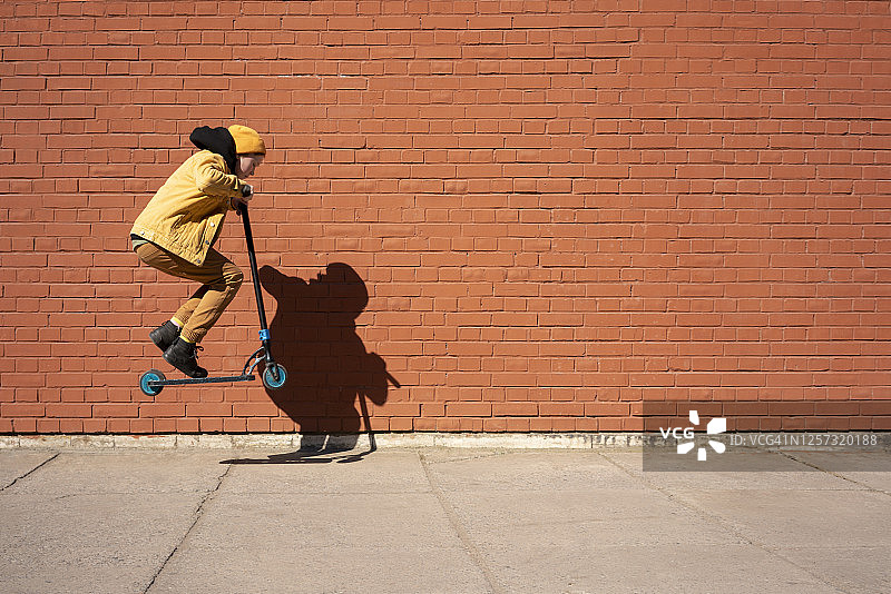 阳光明媚的日子里，男孩在人行道上靠砖墙用滑板车表演特技图片素材