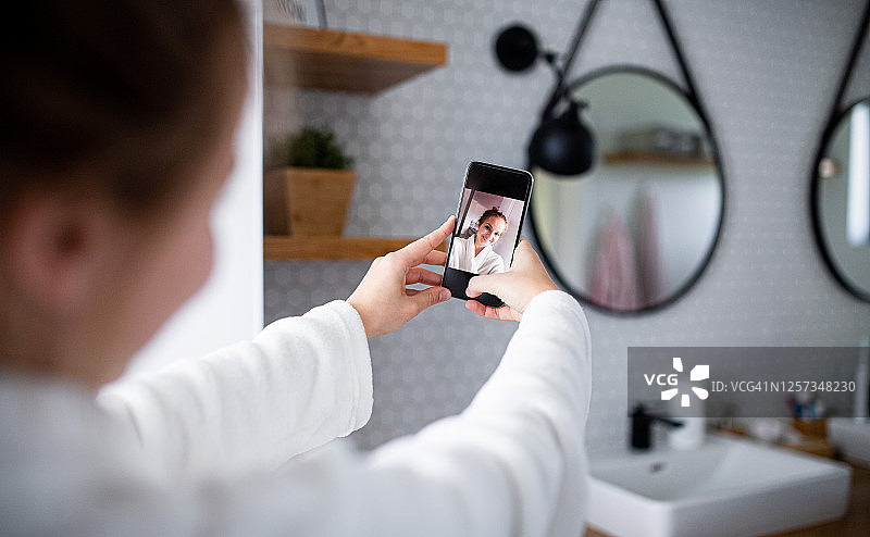 年轻女子穿着浴袍在浴室里用智能手机自拍图片素材