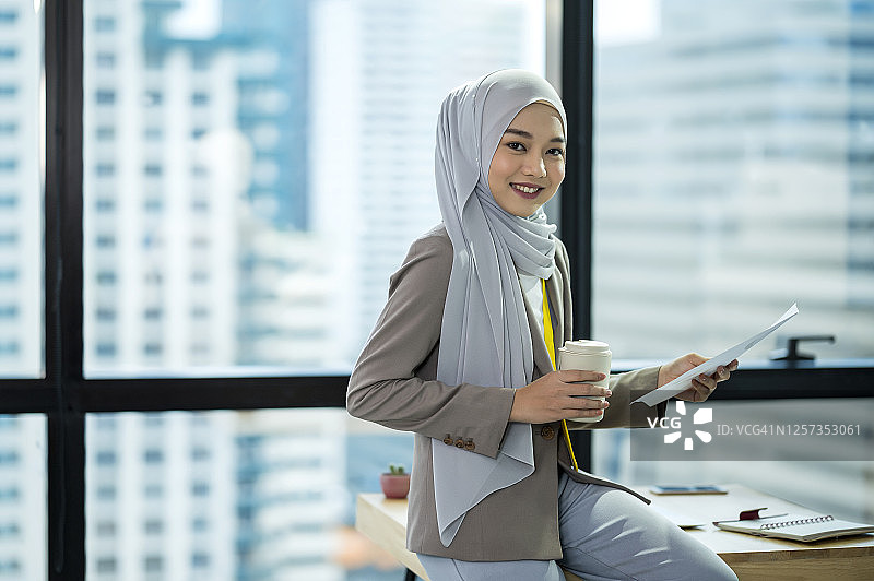 年轻的穆斯林女商人坐在办公室桌子上，在会议室里拿着咖啡杯的肖像图片素材