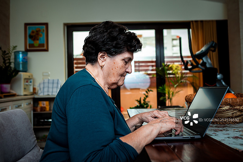 老年女士坐在餐桌旁使用电脑，正在浏览互联网信息。图片素材