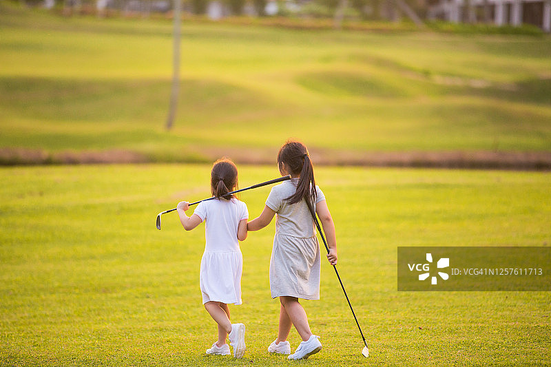 两名亚洲中国儿童走在高尔夫球场上，在美丽的日落时微笑，他们拿着高尔夫球杆图片素材