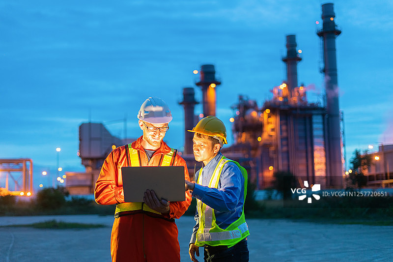 两位工程师戴着安全帽使用笔记本电脑，背景是炼油厂，能源行业的投资合作，日出时分的天空背景下的炼油厂图片素材
