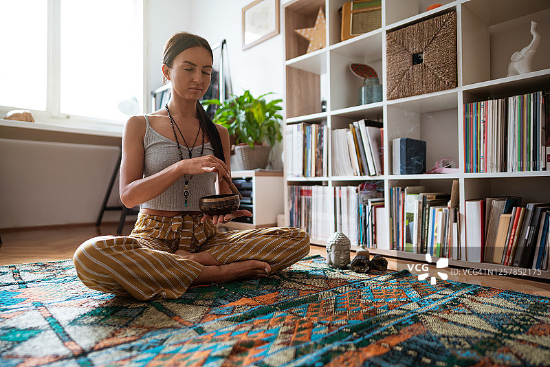 年轻女子闭着眼睛，在家中以莲花坐姿使用冥想铃放松身心图片素材