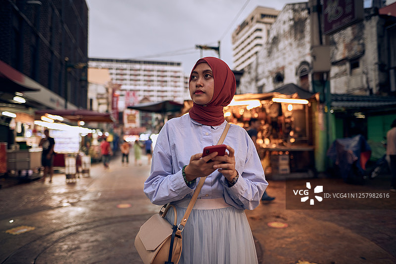 在街头市场户外使用智能手机导航的年轻穆斯林亚洲女性，戴着头巾图片素材