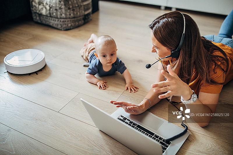 美丽母亲在家用笔记本电脑工作，扫地机器人清洁地面图片素材