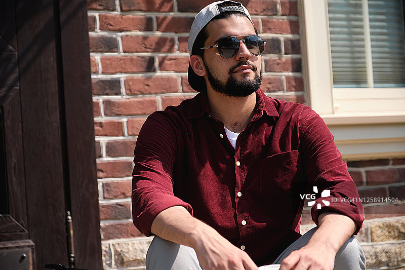 戴着太阳镜、表情严肃的年轻游客肖像。一位戴着棒球帽的深色皮肤印度教印度男子靠在房子的砖墙和木门上。图片素材