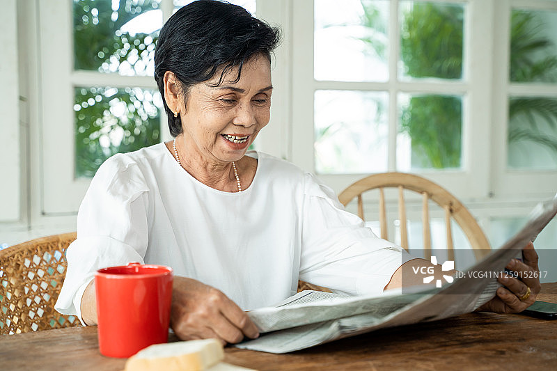 快乐的亚洲老年妇女穿着休闲服在家吃早餐时阅读报纸上的有趣文章。老年妇女享受退休生活方式图片素材
