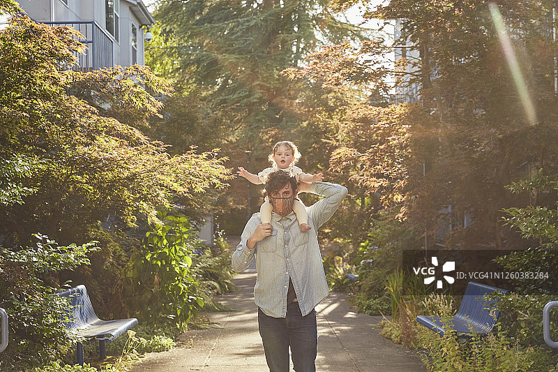 年轻父亲在傍晚带着坐在肩膀上的宝宝散步，戴着防护口罩图片素材