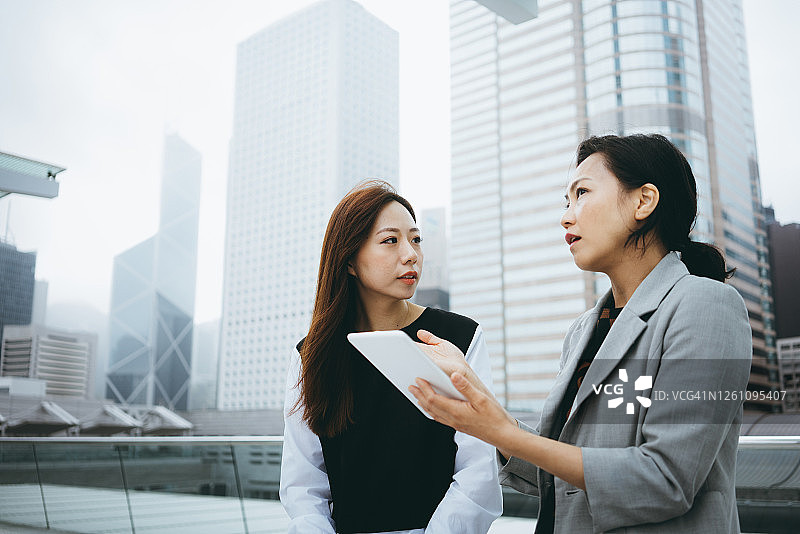 两名女商人在户外使用平板电脑讨论商业项目，背景是城市景观和现代办公楼图片素材