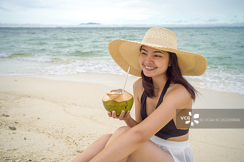 晴朗天气下坐在海滩边喝椰子汁的亚洲女性图片素材