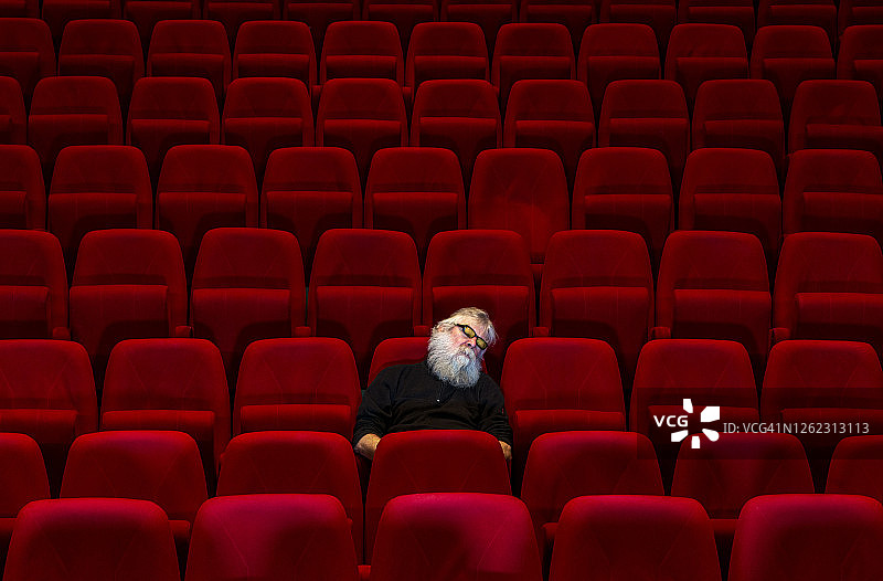 一名白胡子男人在空旷的电影院或剧院里舒适的红色座位上睡觉图片素材