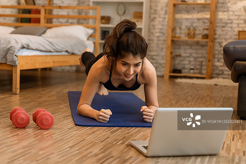 20多岁的亚洲年轻女人穿着运动服，在电脑笔记本上观看健身训练课程，做平板支撑。健康的女孩在卧室里锻炼和学习。网络教育理念图片素材