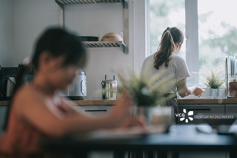 女儿在家学习，在餐厅写中国书法，母亲在厨房准备食物图片素材