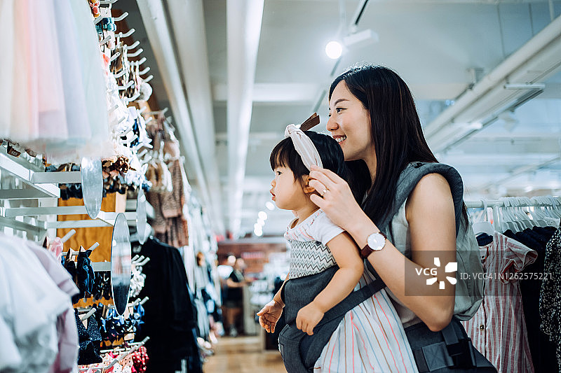 年轻的亚洲母亲在服装店购物时，在镜子前为小女儿戴头带图片素材