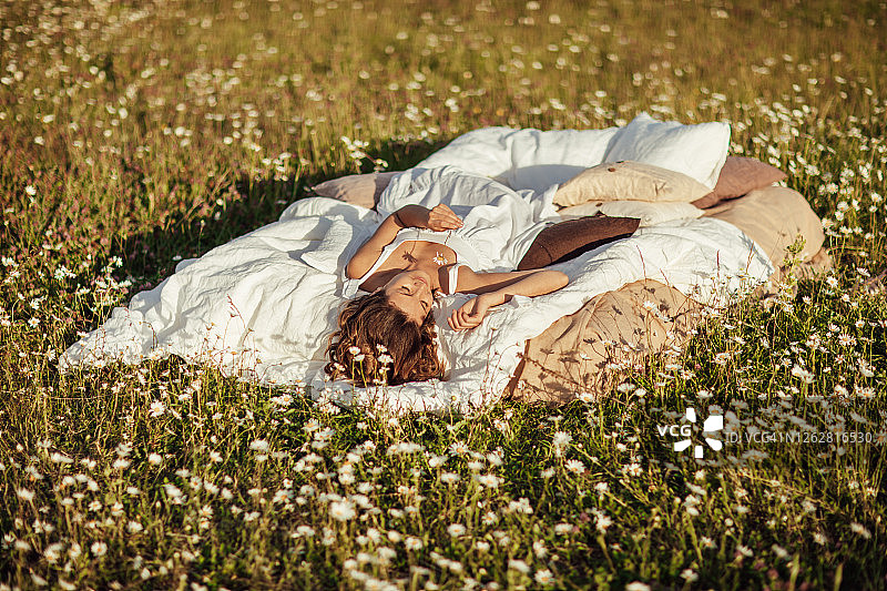 可爱的年轻卷发女人穿着白色连衣裙，躺在夏季洋甘菊田里柔软的亚麻床上。夏季假期灵感，良好睡眠和放松的概念图片素材