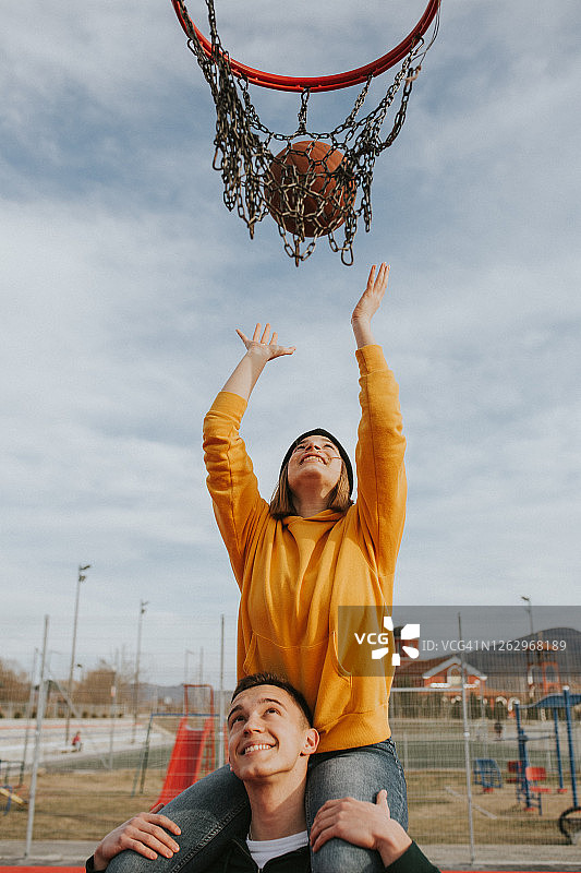 女朋友坐在男朋友的肩膀上在篮球场上投篮图片素材