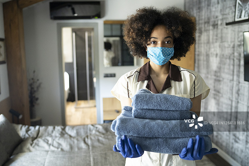 年轻的女保洁员拿着毛巾在酒店房间里，戴着面罩图片素材